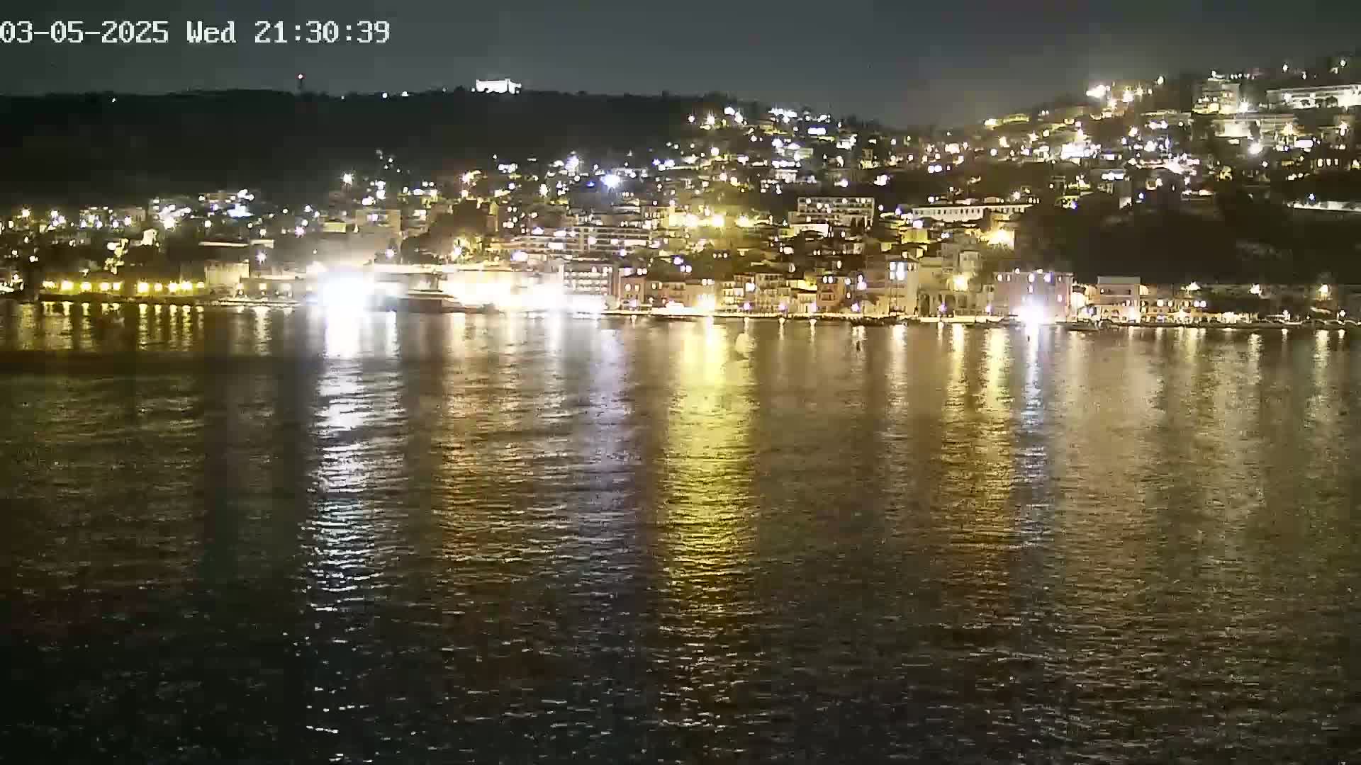 A nighttime view of a town's waterfront, illuminated by lights reflecting on calm water.