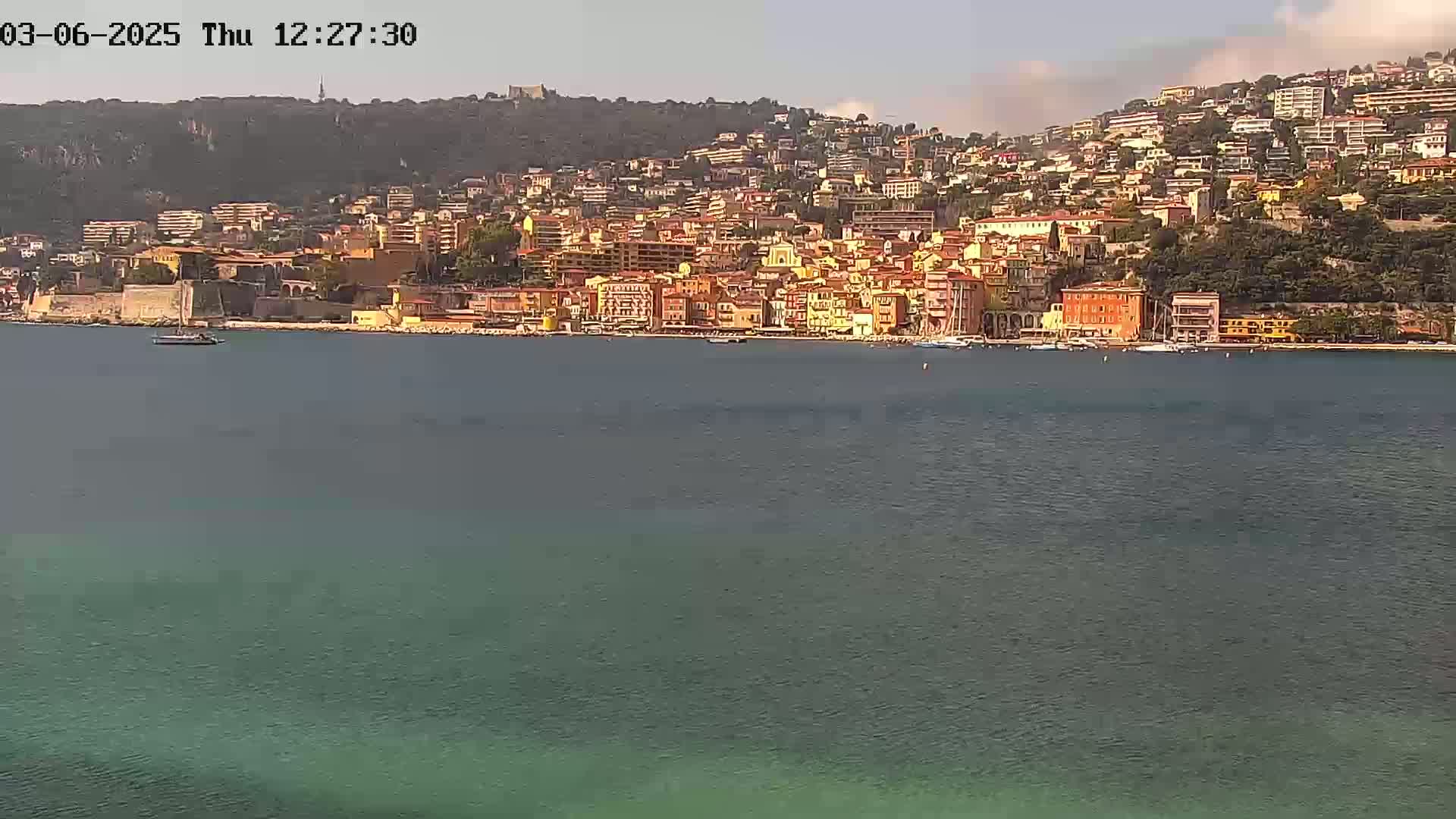 A coastal town with colorful buildings situated on a hillside overlooking calm, greenish-blue water under a mostly clear sky.