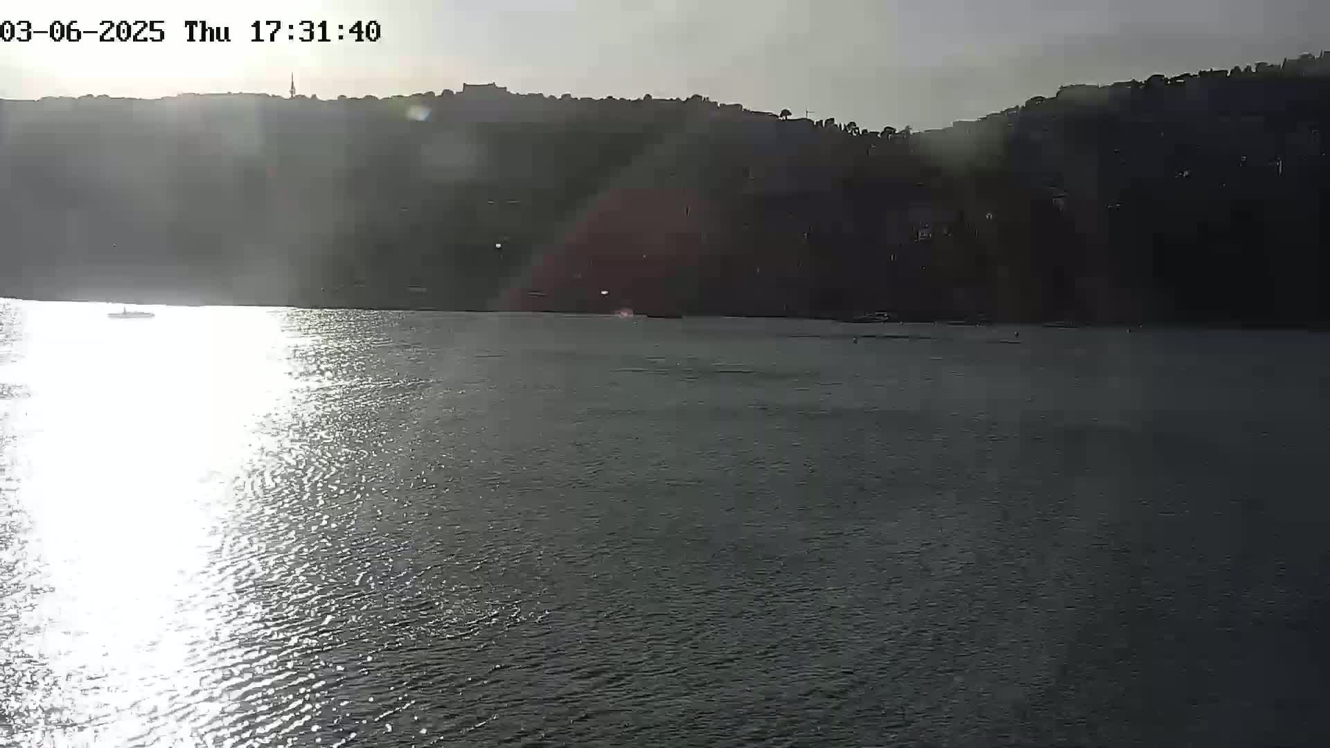 A body of water reflects sunlight, with a distant shoreline and hills visible under a partly sunny sky.