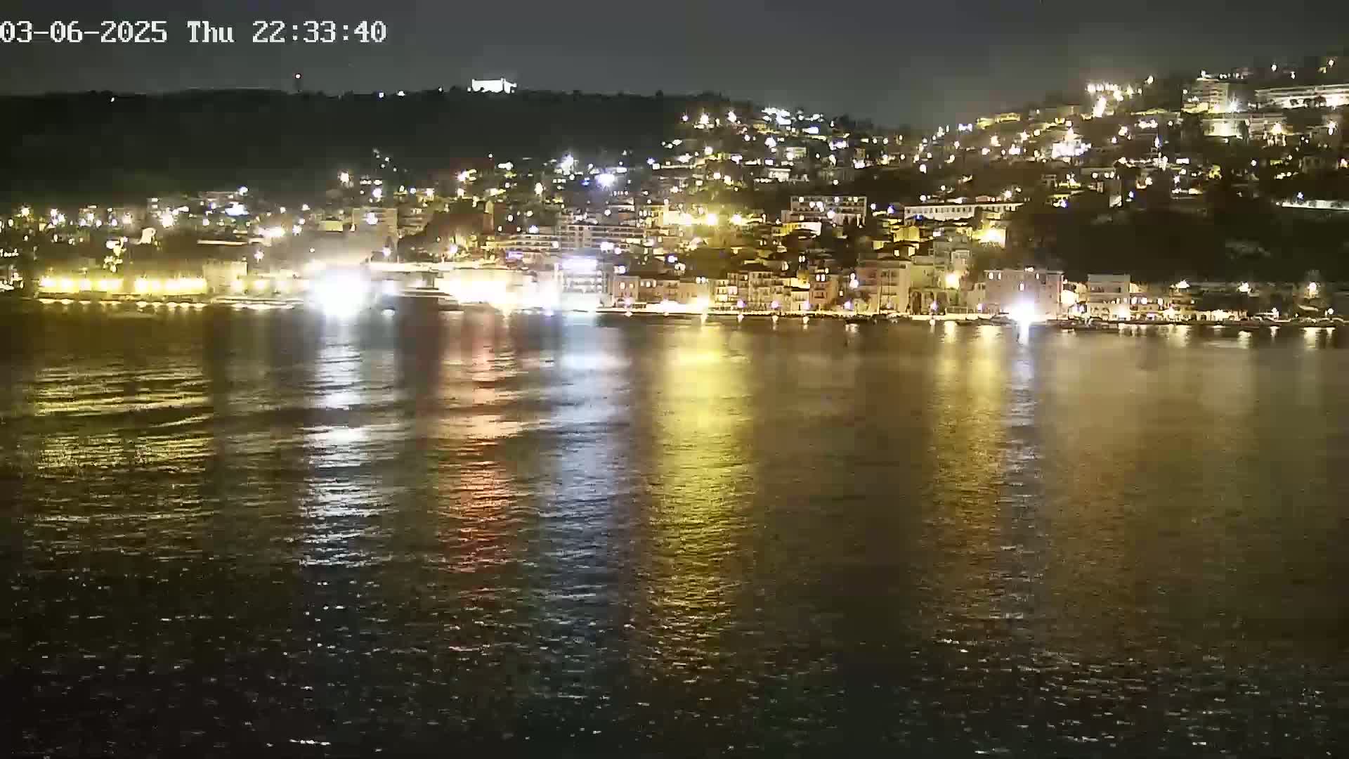 A nighttime view of a brightly lit town situated on a hillside overlooking a calm body of water.