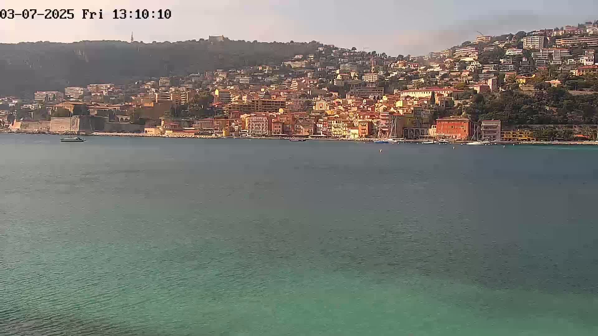 A coastal town with colorful buildings climbs a hillside, overlooking a calm, teal-colored sea on a sunny day.