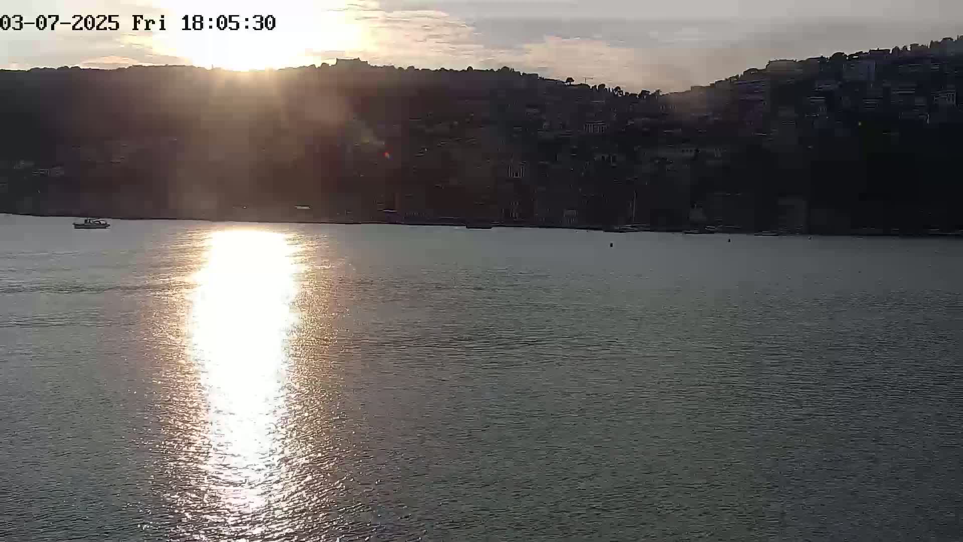 A calm body of water reflects the setting sun, with a hillside town visible in the background under a partly cloudy sky.
