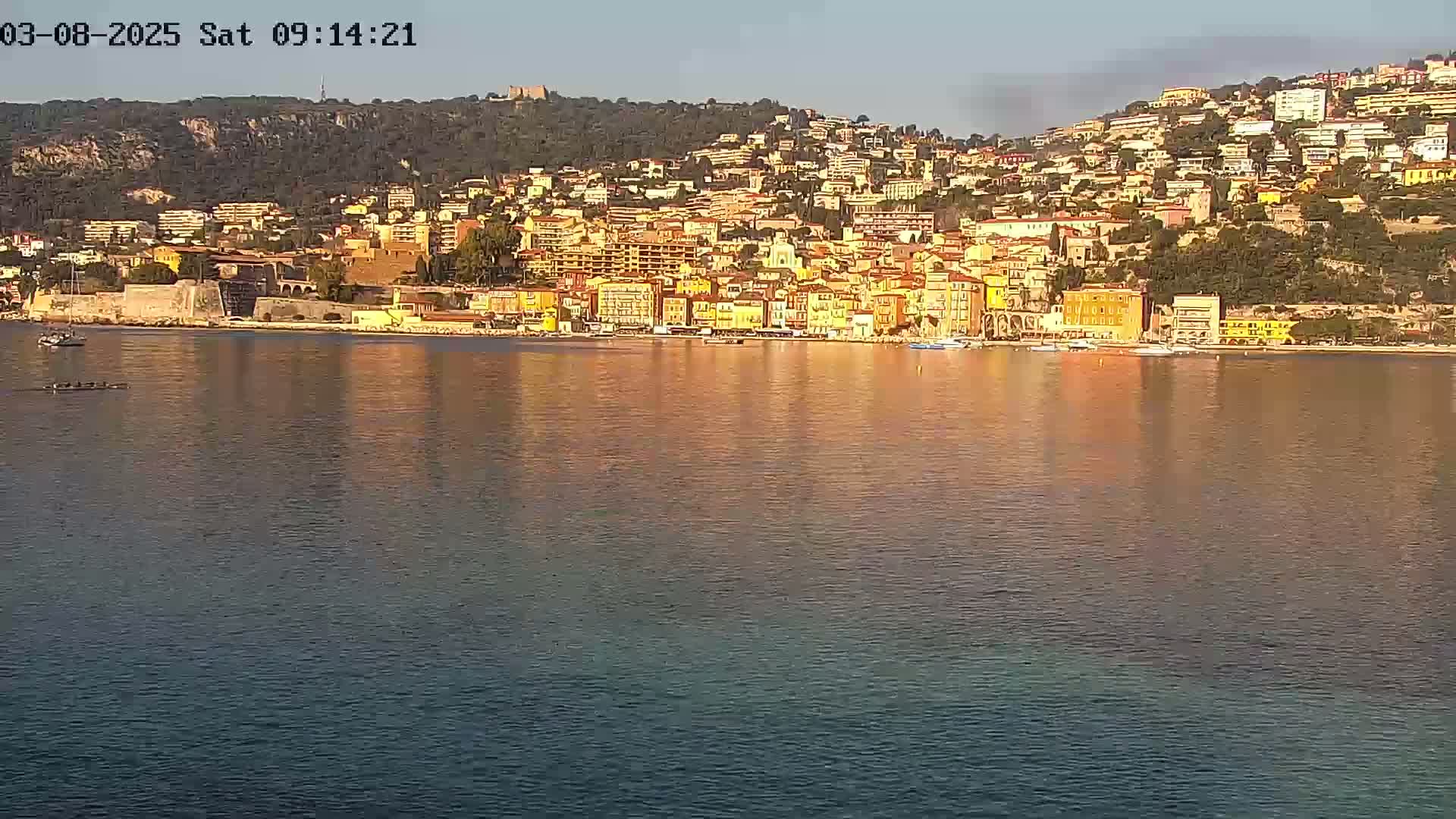 A coastal town of multi-story buildings in warm colors sits on a hillside overlooking calm water under a clear sky.