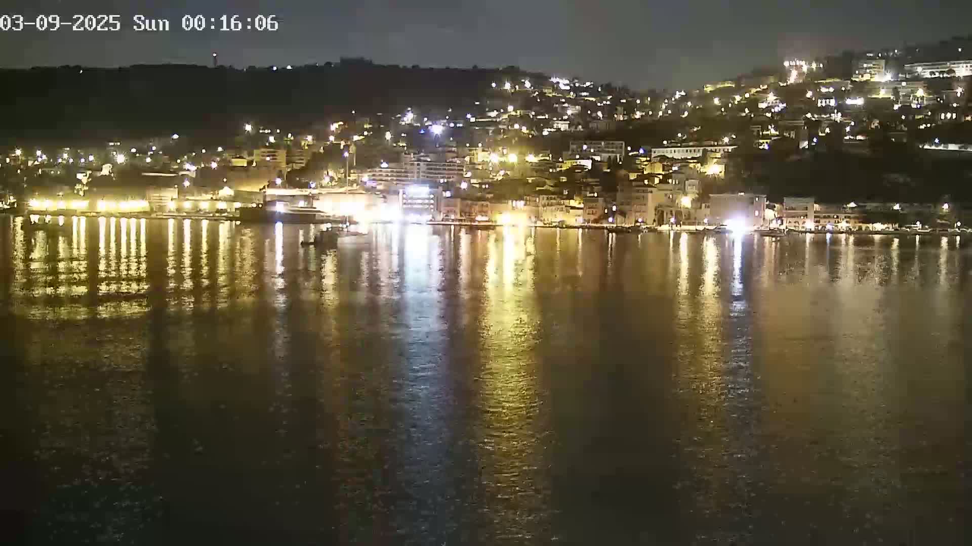 A nighttime view of a harbor town on a calm body of water, with the lights of buildings reflecting on the surface.