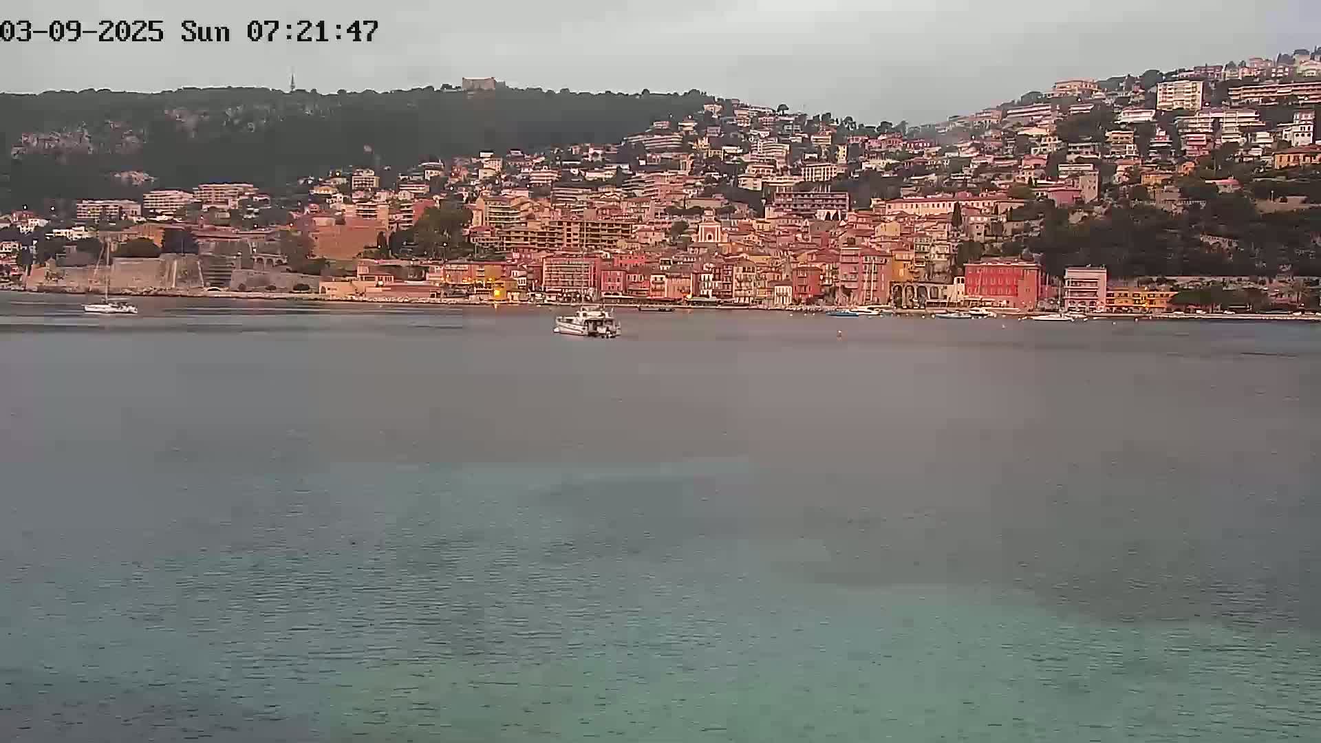 A small boat is in the calm water in front of a hillside town under an overcast sky.