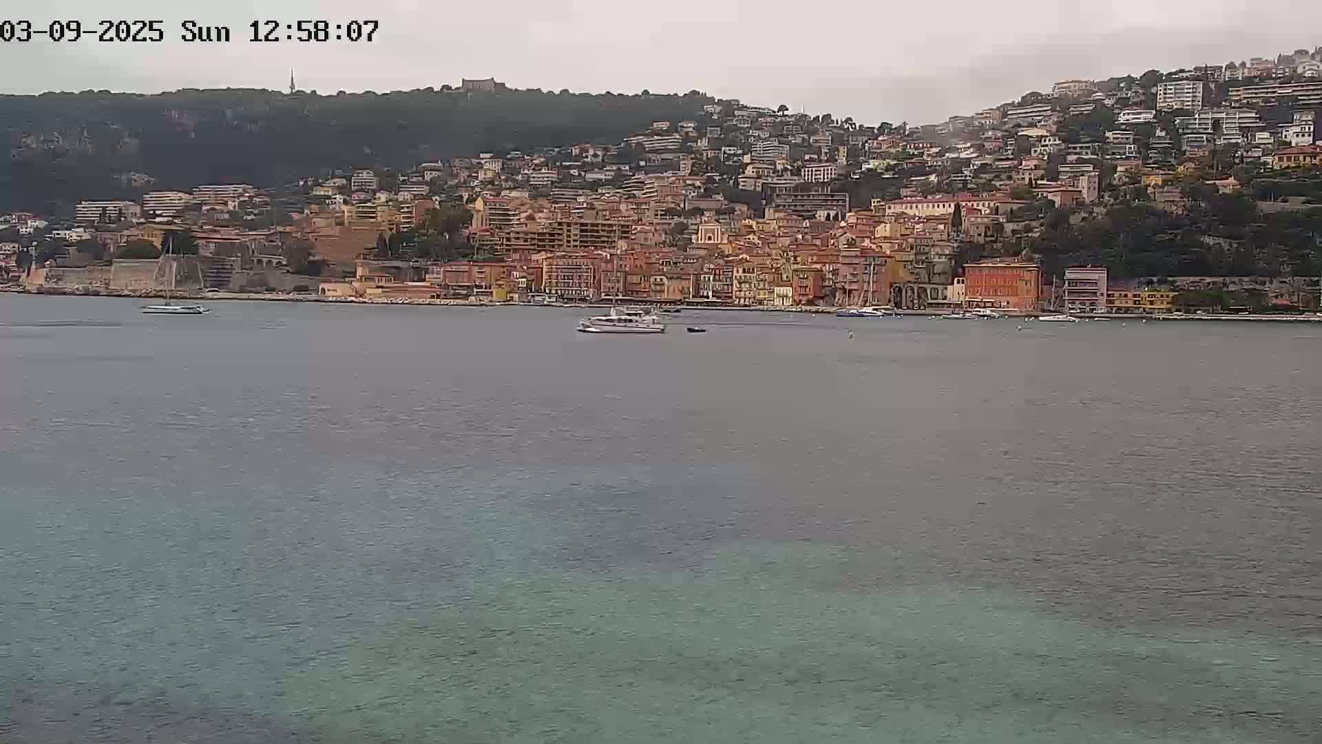 A coastal town with many buildings climbs a hillside overlooking a calm body of water under an overcast sky.