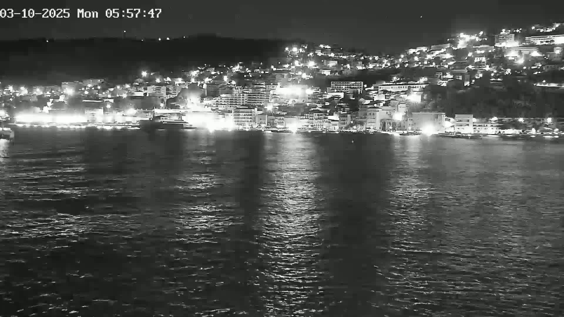 A nighttime, black and white view of a city's waterfront at night, illuminated with lights, across a body of dark water.