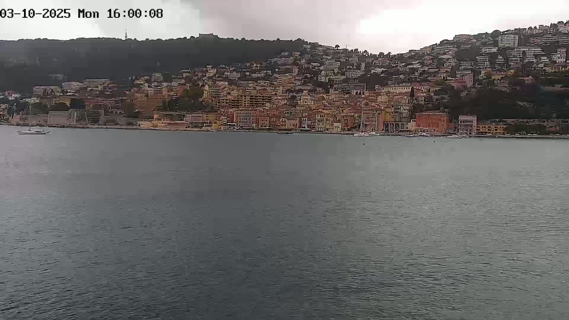 A coastal town with many buildings of varying heights sits on a hillside overlooking a calm body of water under an overcast sky.