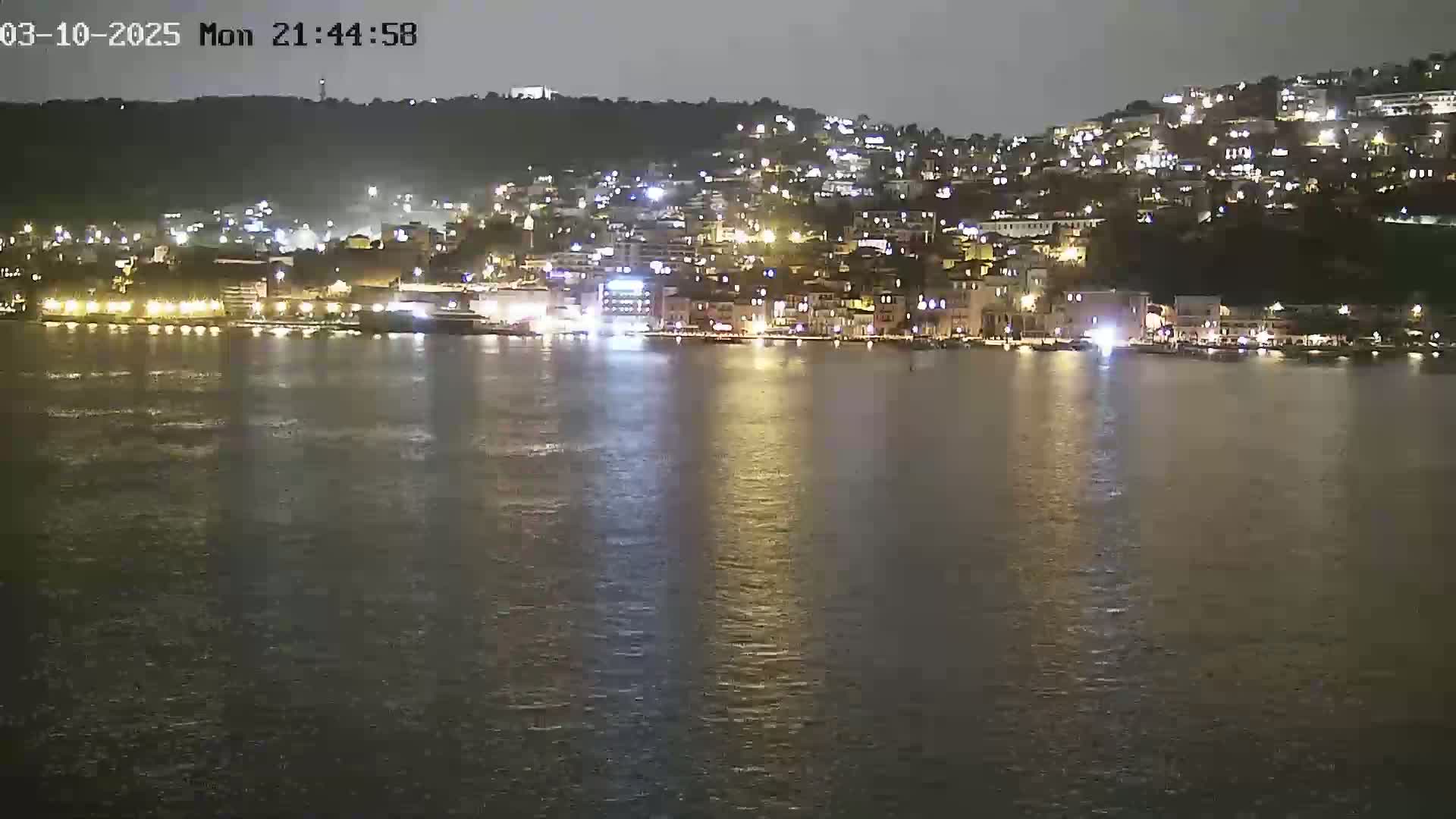 A nighttime view of a city illuminated along a calm body of water, nestled against a dark hillside.