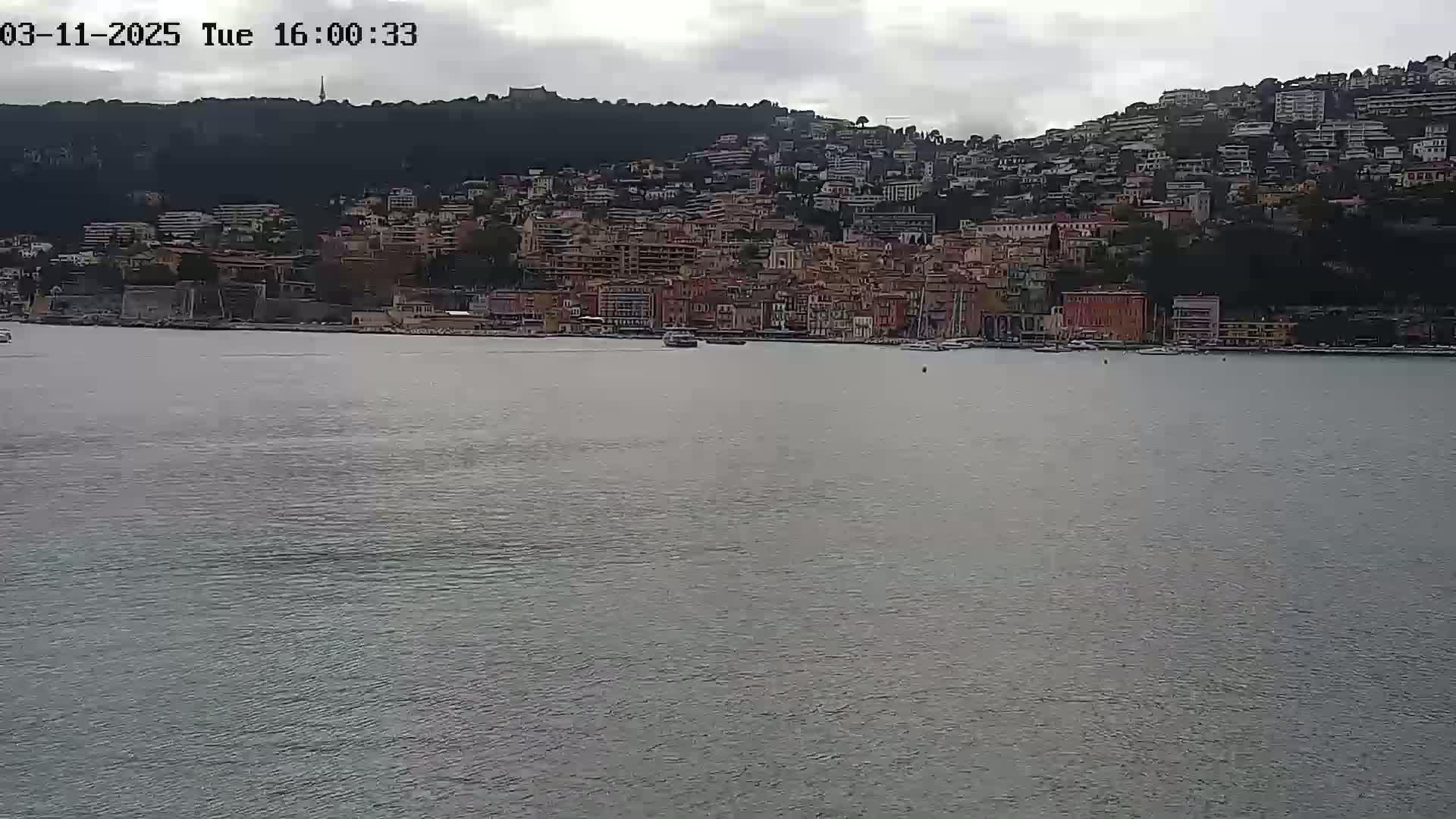 A coastal town with many buildings climbs a hillside overlooking a calm body of water under an overcast sky.
