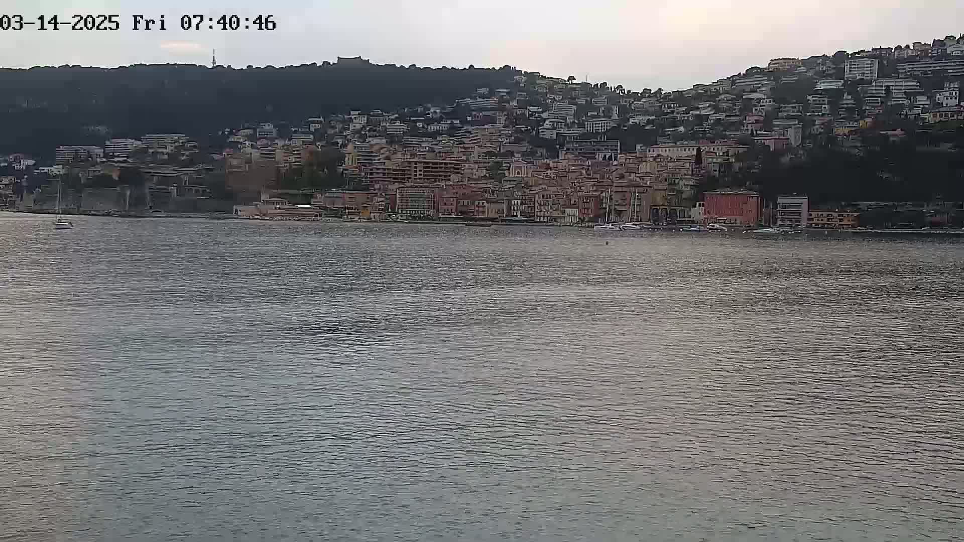 A coastal town densely packed with buildings sits on a hillside overlooking calm water under an overcast sky.