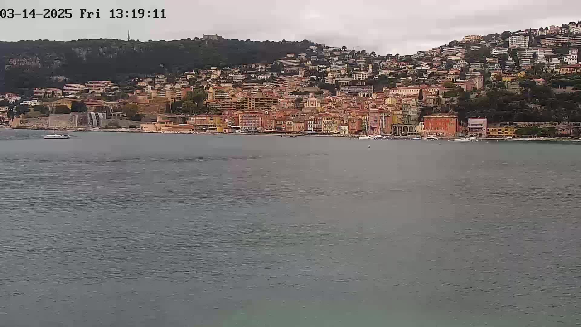 A coastal town with many buildings of varying heights and colors sits on a hillside overlooking a calm body of water under an overcast sky.