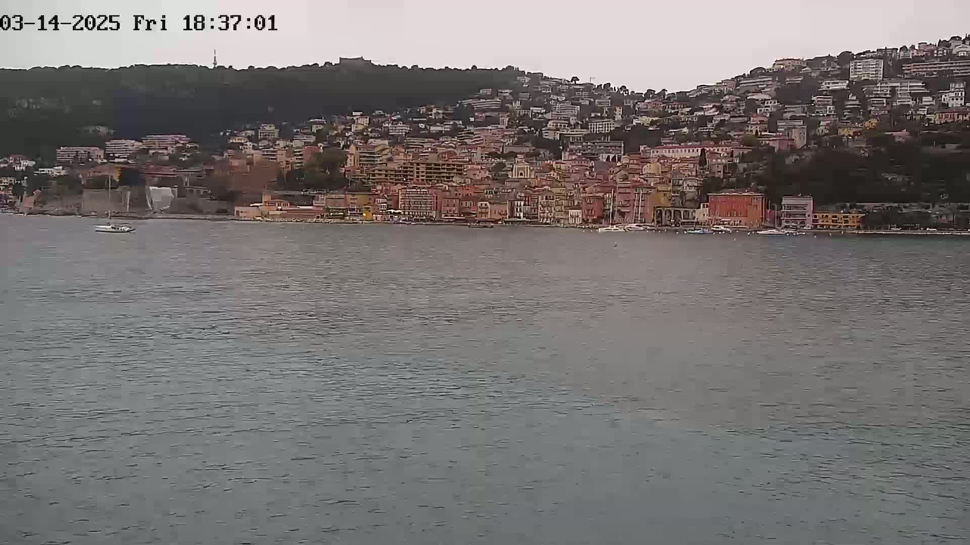 A coastal town with numerous buildings on a hillside is visible across a calm body of water under an overcast sky.