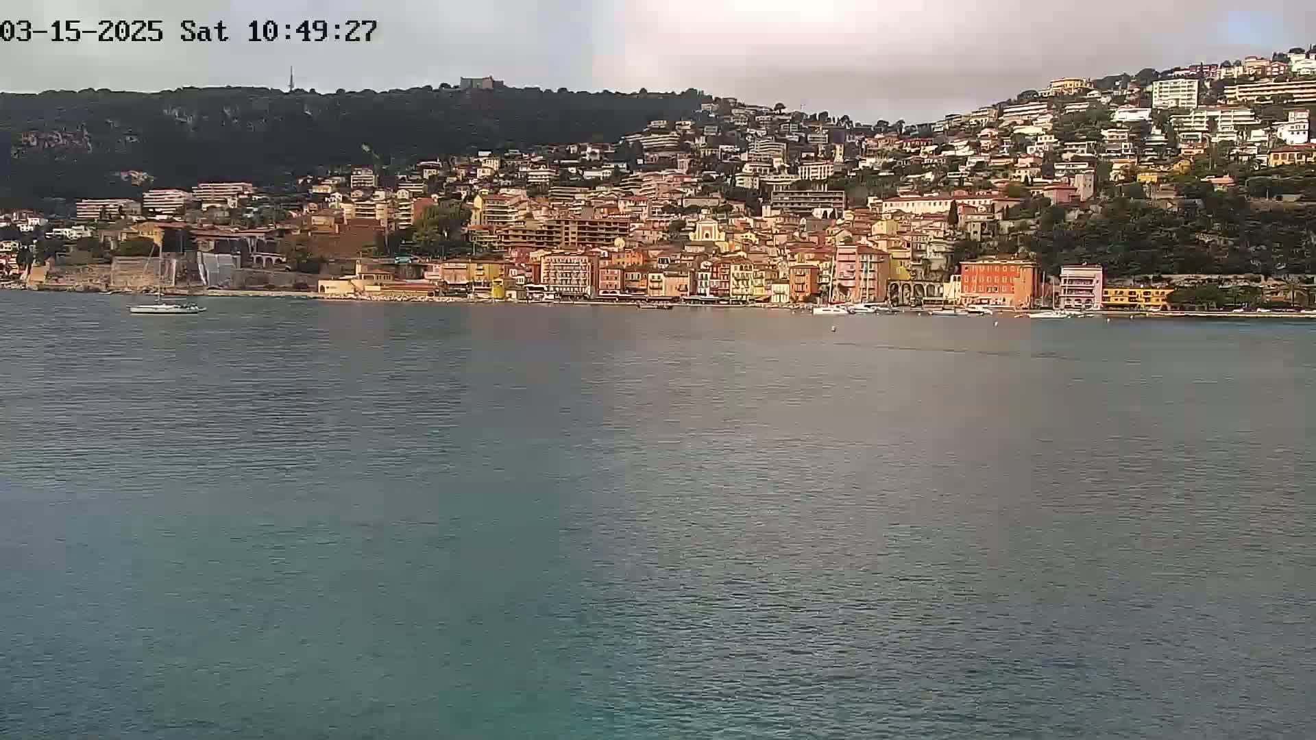 A colorful waterfront town, nestled against a hillside, is seen across a calm body of water under an overcast sky.