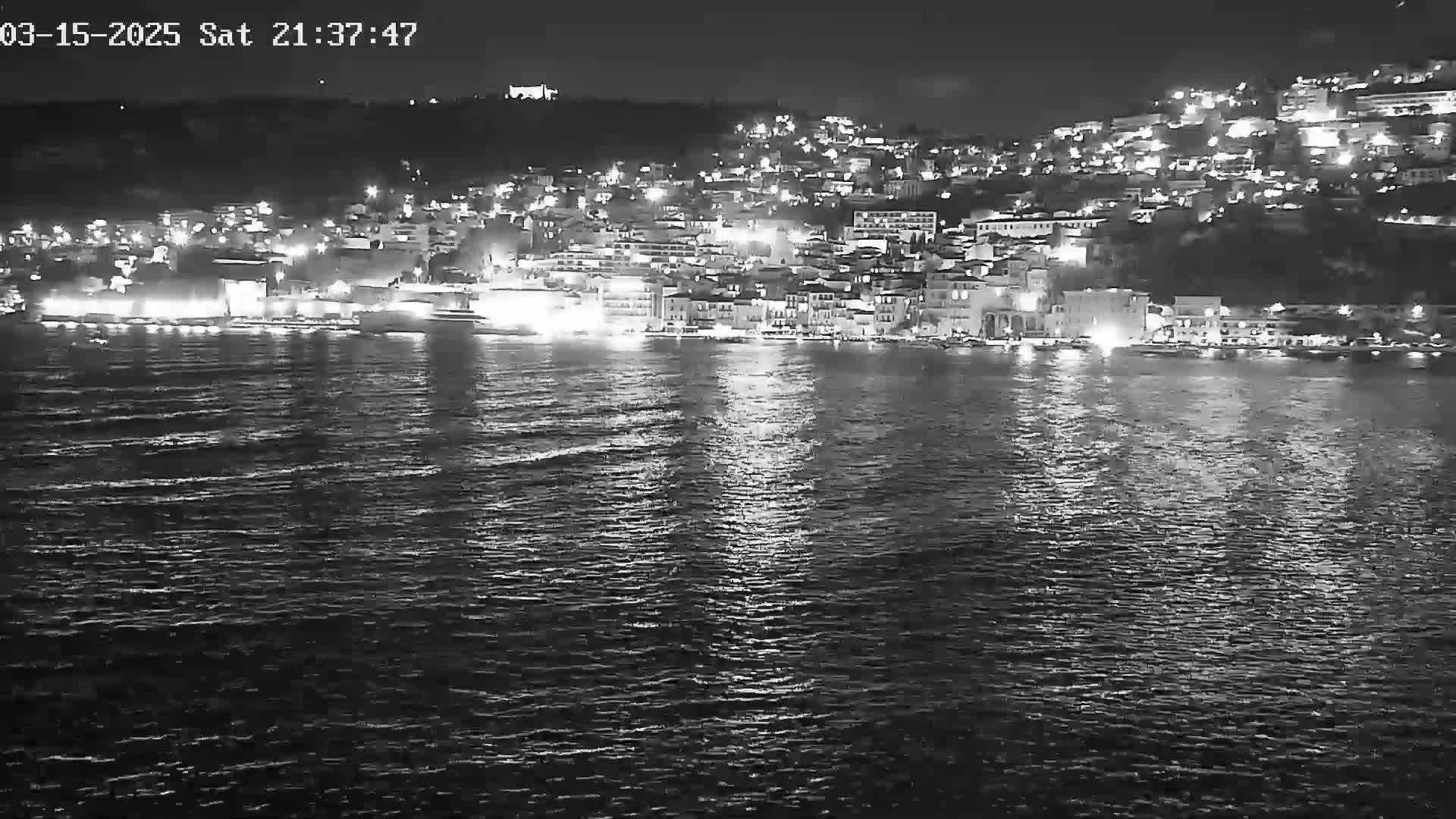 A nighttime view of a city's illuminated waterfront, situated on a hillside, reflected on dark, gently rippling water.