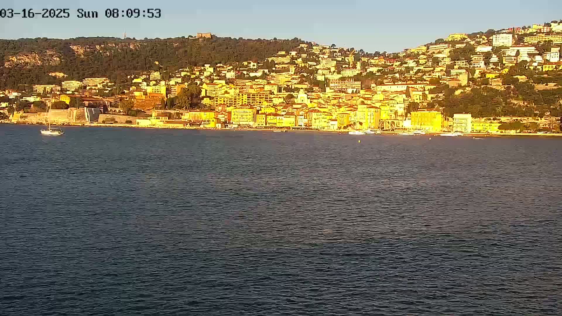 A small boat is visible on calm water in front of a hillside town under a clear sky.