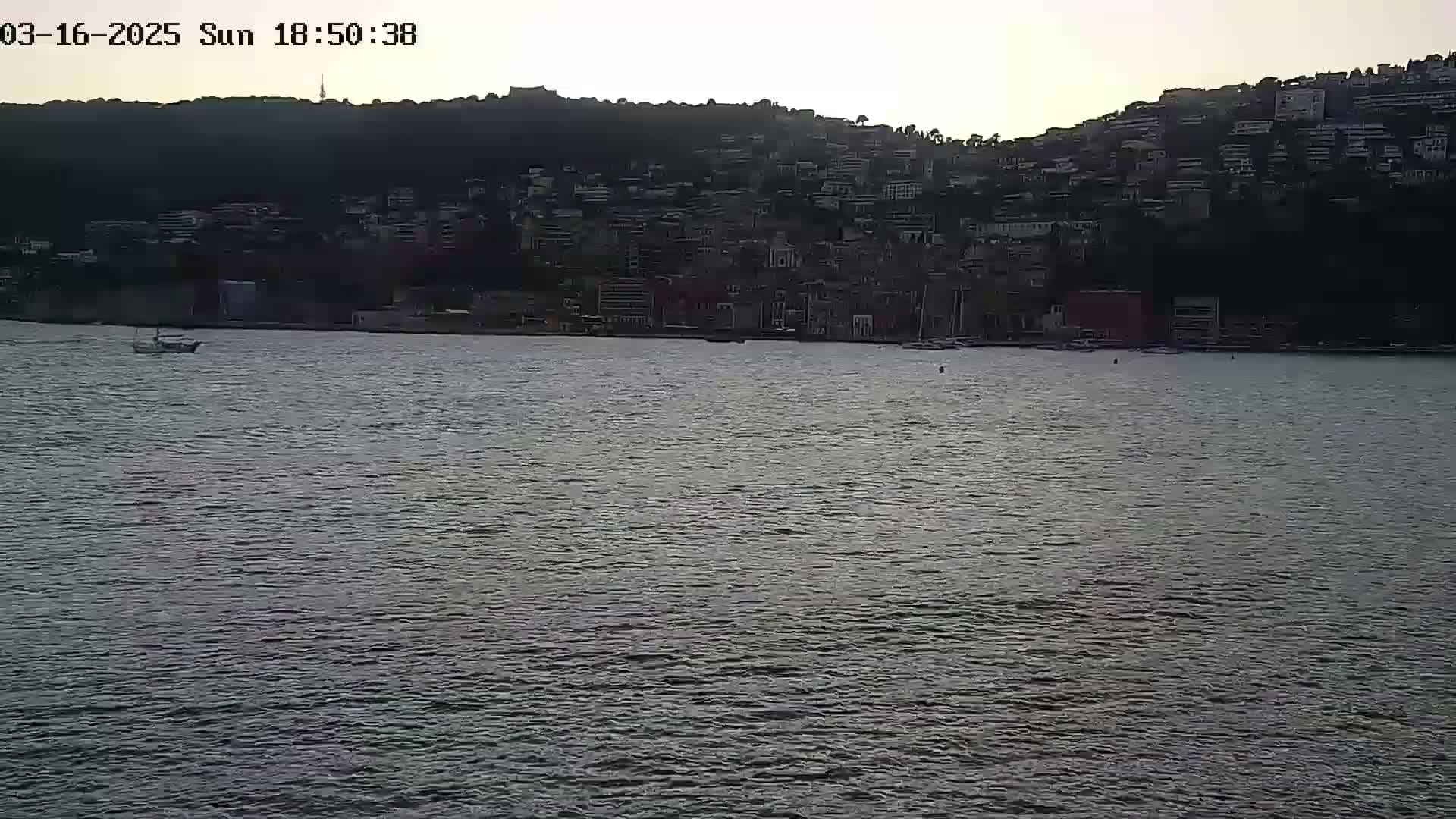 A small boat is on calm water in front of a hillside town at dusk.