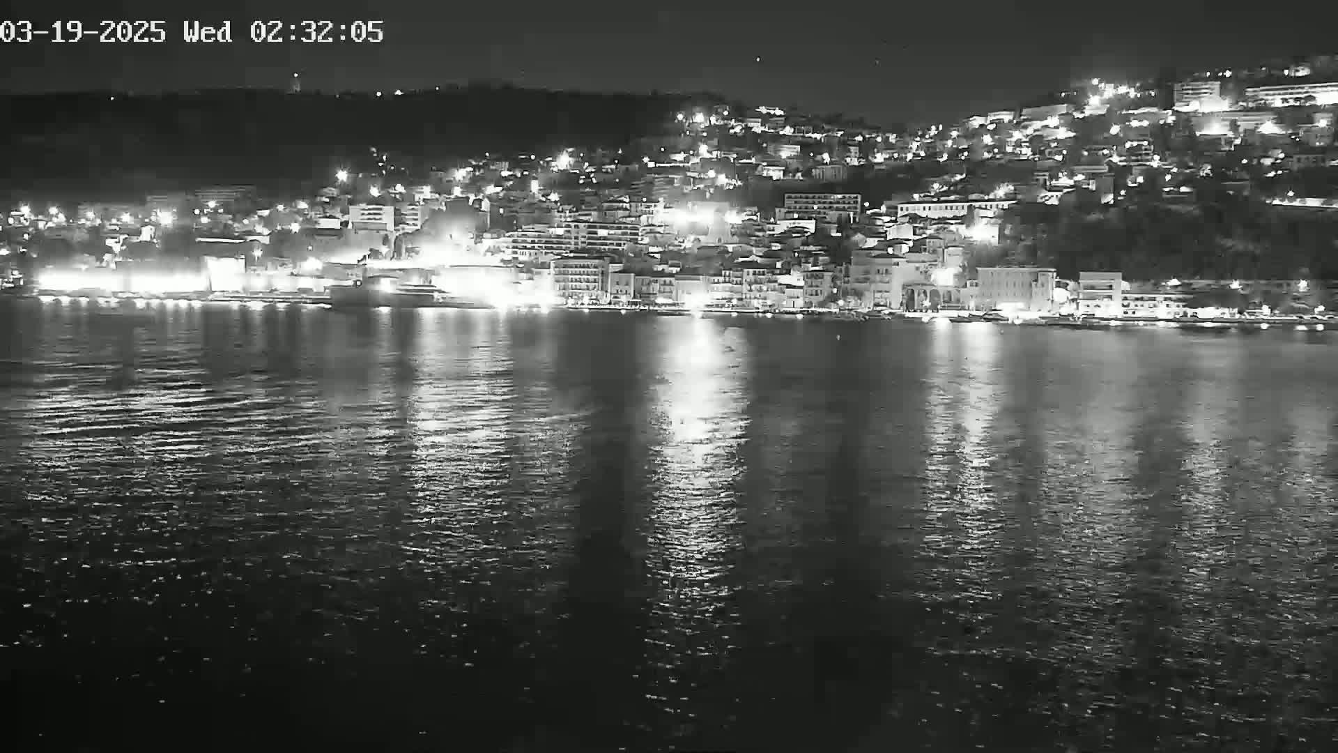 A nighttime view of a city's waterfront, illuminated by lights reflecting on calm water.