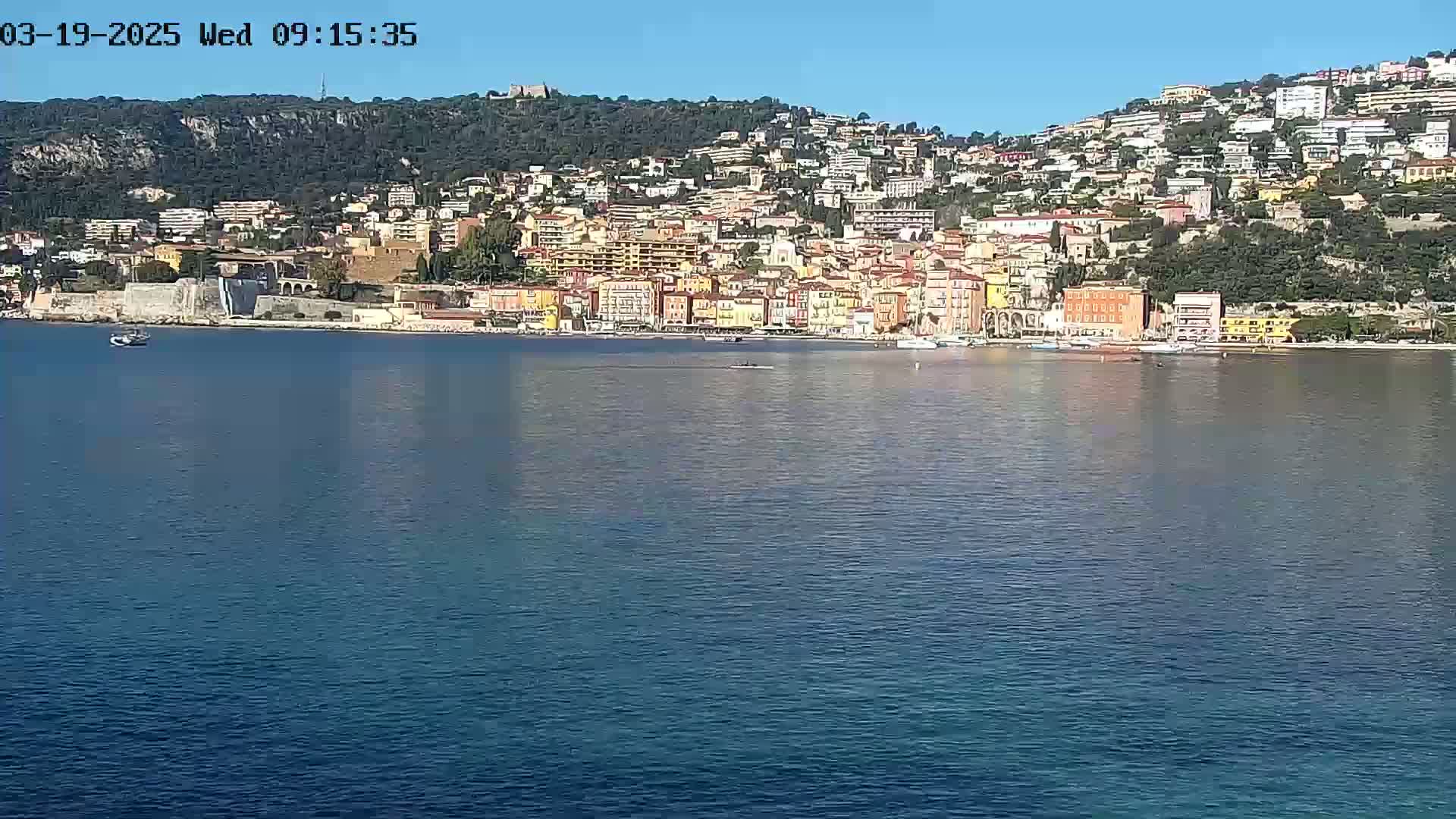 A coastal town with numerous buildings climbs a hillside overlooking a calm, blue body of water under a clear sky.