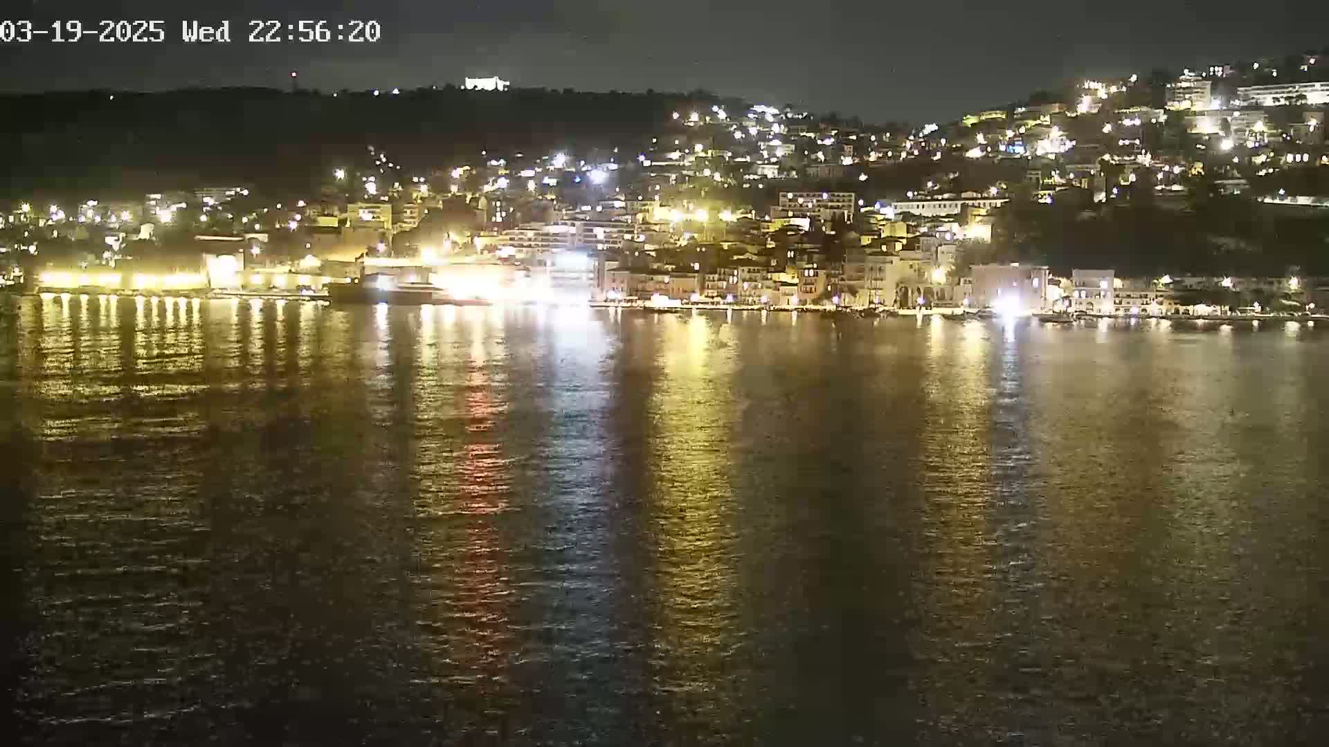 A nighttime view of a city on a hillside, brightly lit, with its lights reflecting on calm water.