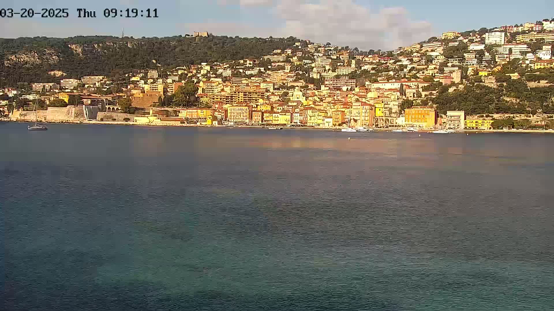 A coastal town with colorful buildings climbs a hillside overlooking a calm, dark-blue body of water under a partly cloudy sky.