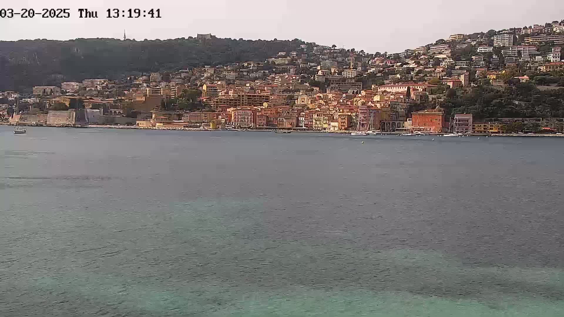 A coastal town with many buildings of varying heights and colors sits on a hillside overlooking calm, greenish-blue water under a mostly clear sky.