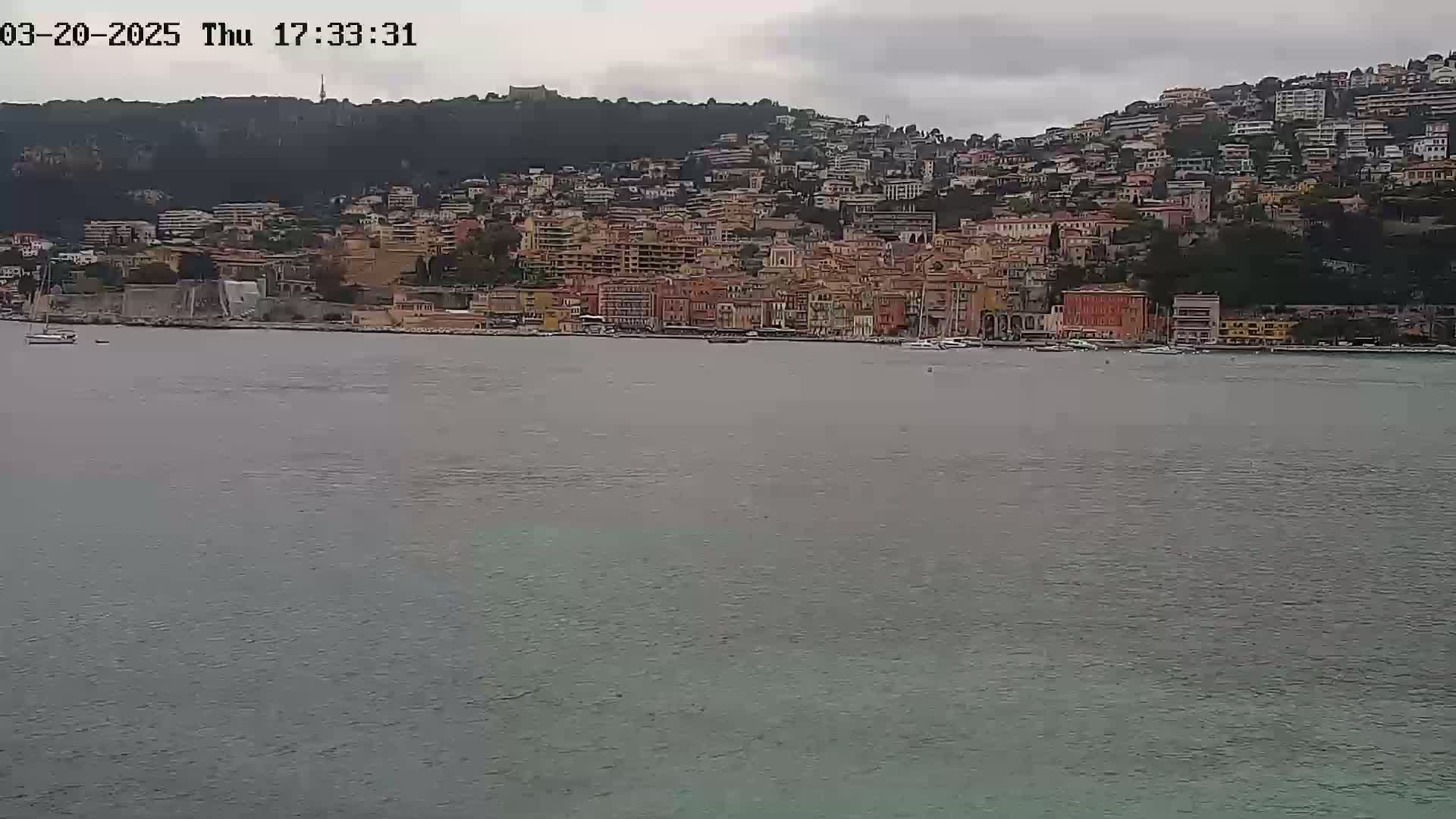 A coastal town with many buildings on a hillside is seen across a calm body of water under an overcast sky.