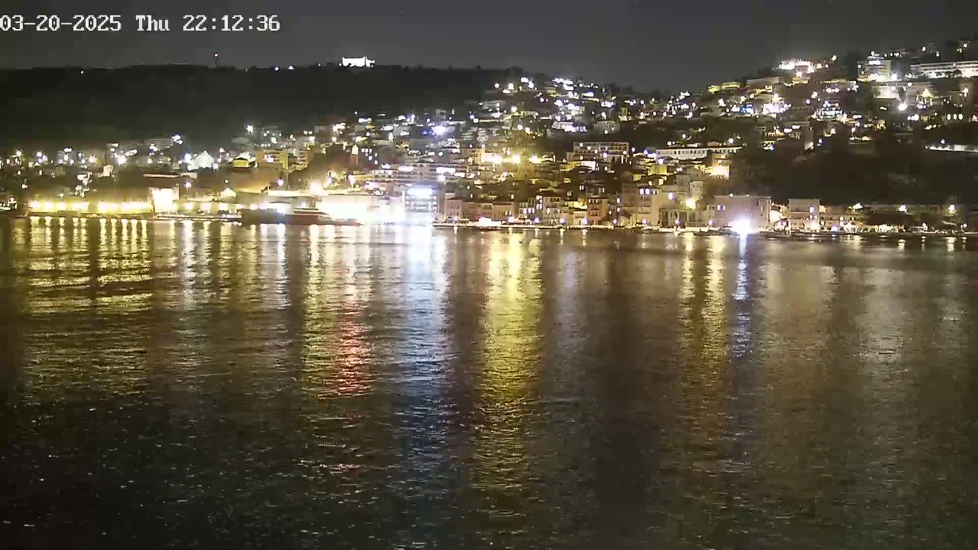 A nighttime view of a brightly lit town nestled on a hillside, its lights reflected in the calm water of a bay.