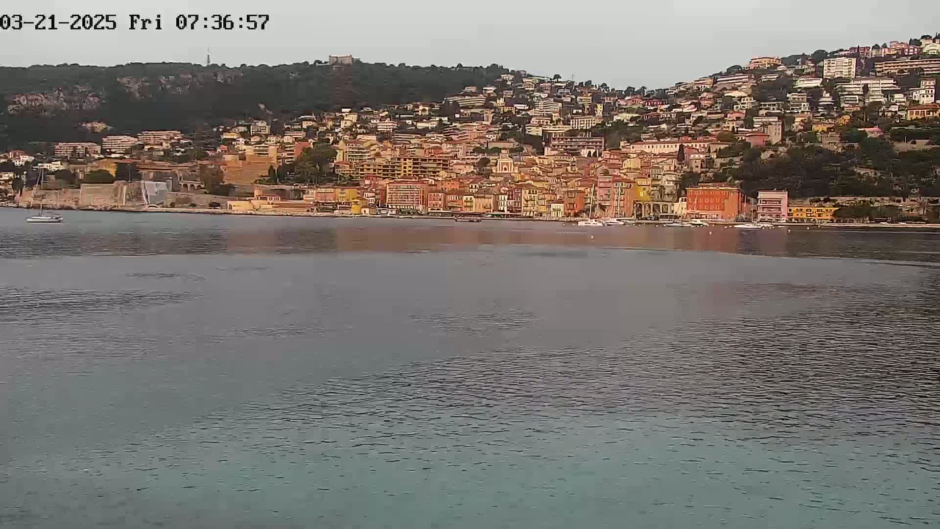 A coastal town with multi-story buildings of various colors sits on a hillside overlooking calm water under an overcast sky.