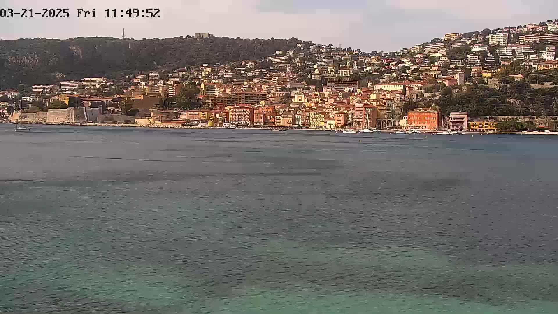 A coastal town with numerous buildings of varying heights, primarily orange and yellow, sits on a hillside overlooking calm, dark-green water under a mostly clear sky.