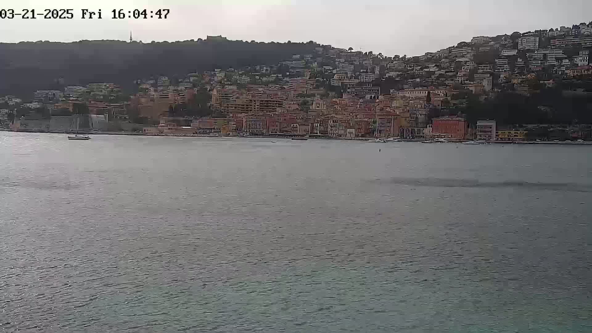 A coastal town with many closely-packed buildings sits on a hillside overlooking a calm body of water under an overcast sky.