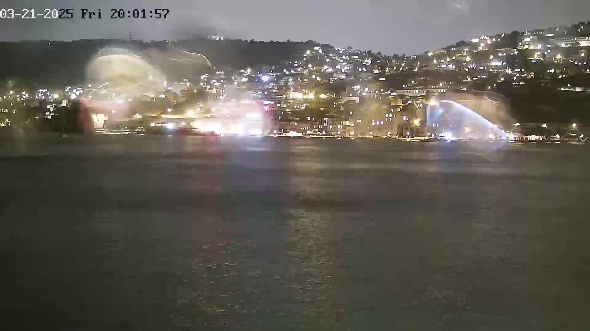 A nighttime view of a city illuminated along a dark body of water, with what appears to be fireworks or water fountains erupting from the waterfront.