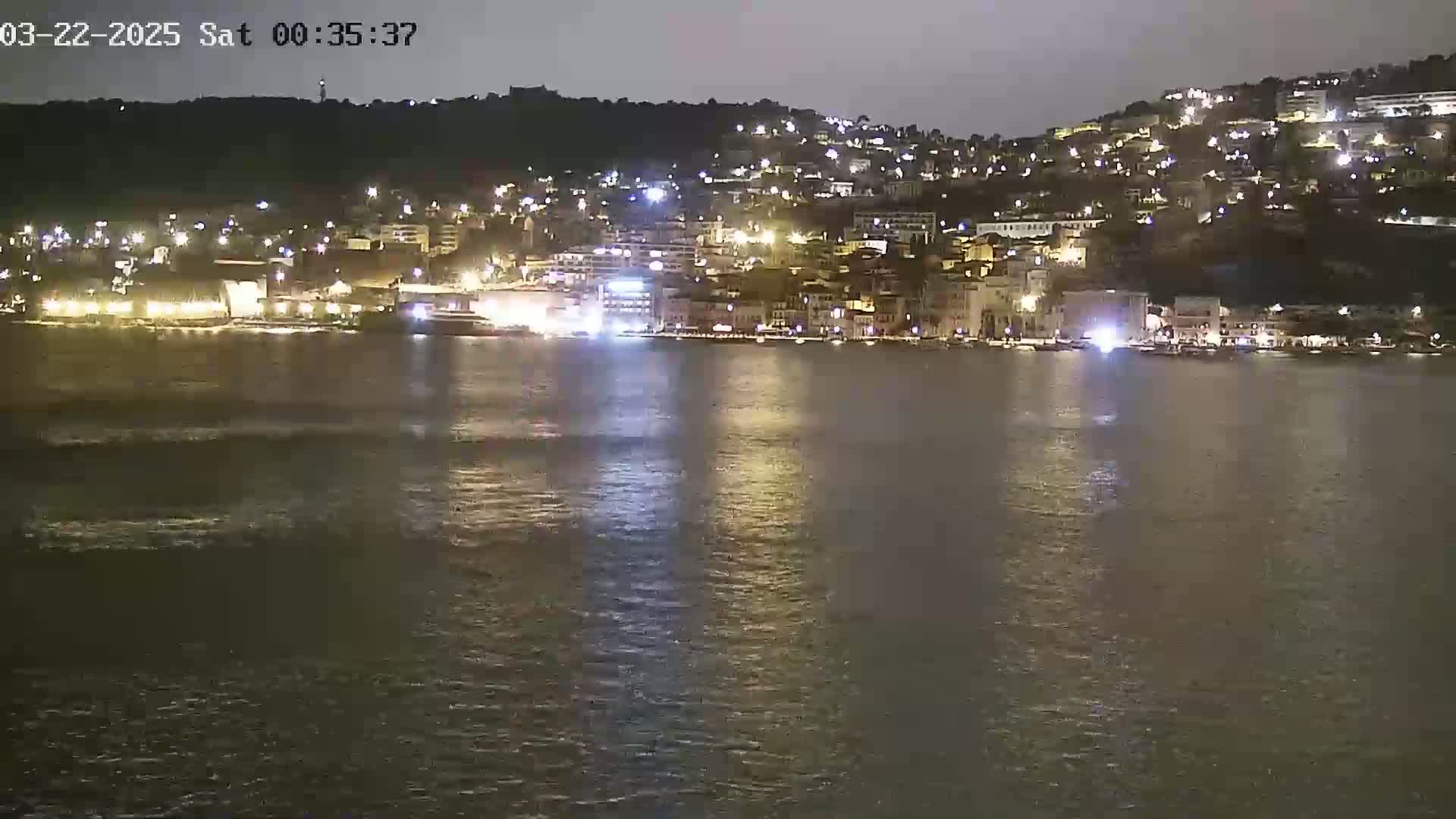 A nighttime view of a city's waterfront, illuminated by many lights, across a calm body of water.