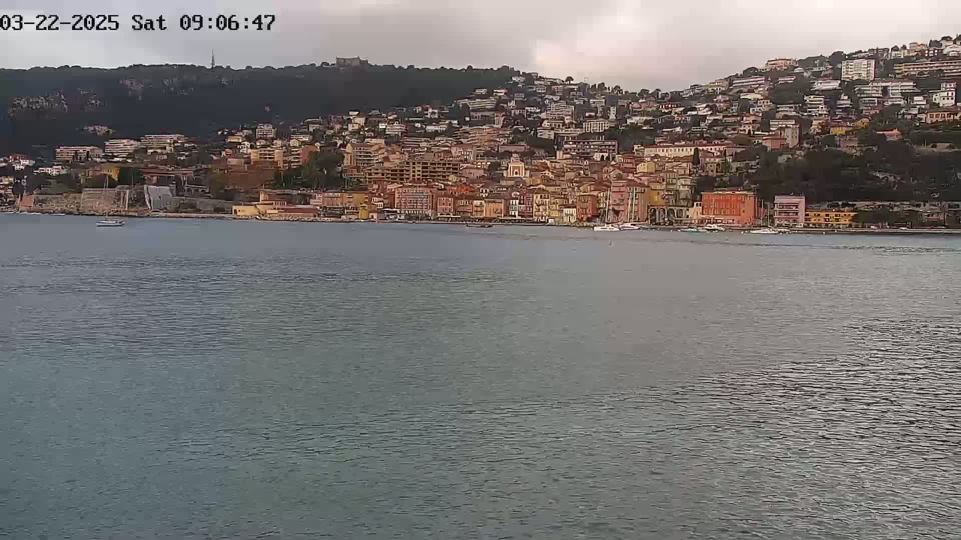 A coastal town with many buildings of varying heights and colors sits on a hillside overlooking a calm body of water under an overcast sky.