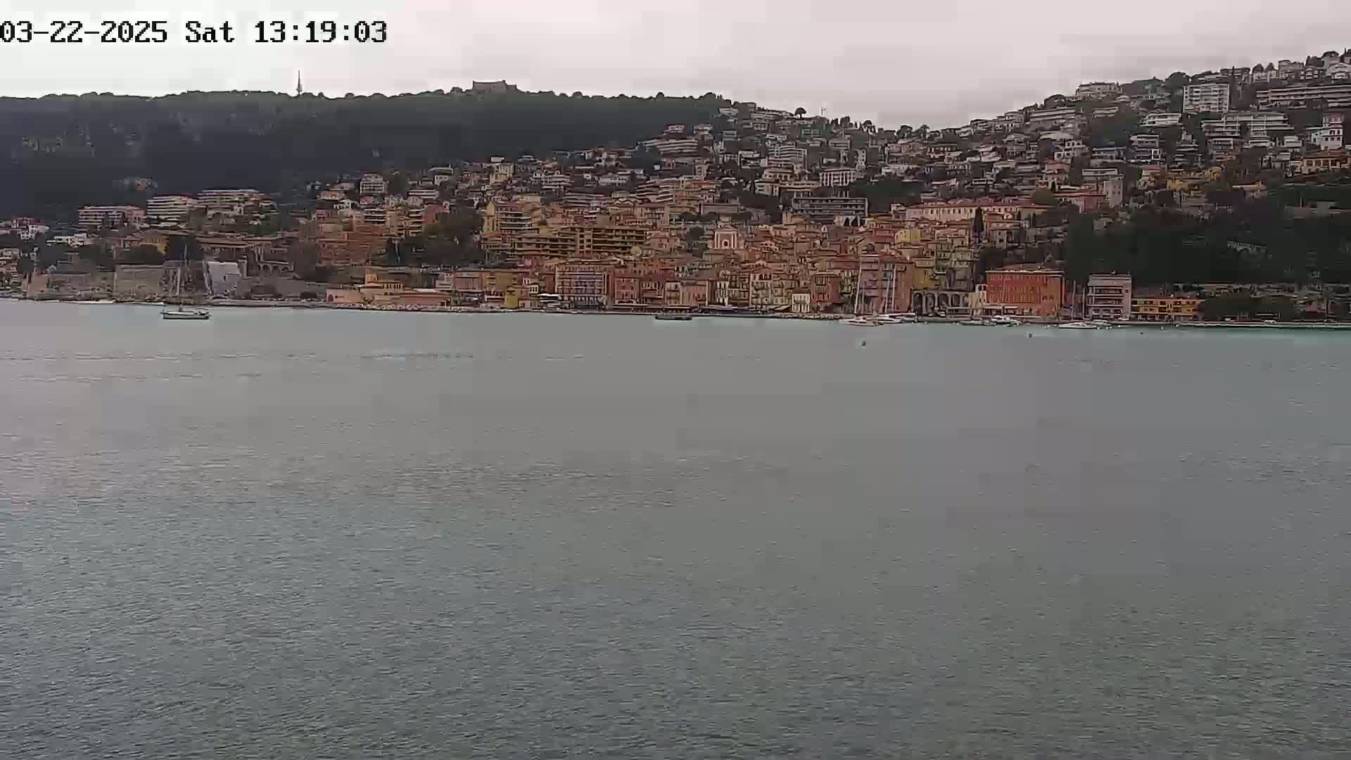 A coastal town with many buildings of varying heights, nestled against a hillside, is viewed from across a calm body of water under an overcast sky.