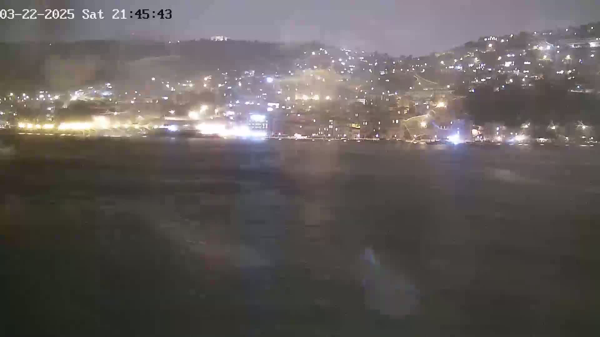 A nighttime view of a city on a hillside across a dark body of water, under cloudy skies.