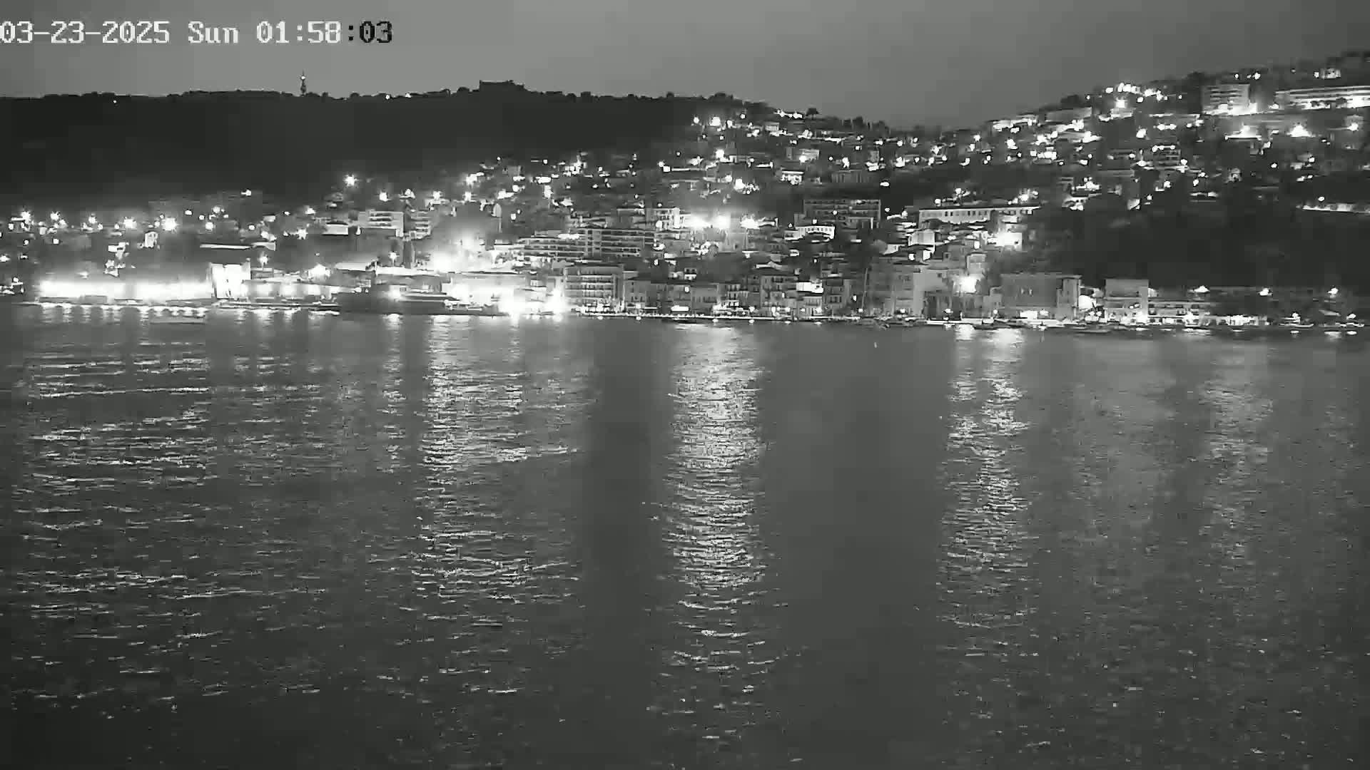 A nighttime, low-light view of a town situated on a hill overlooking a calm body of water, with many lights illuminating the buildings and reflecting on the water's surface.