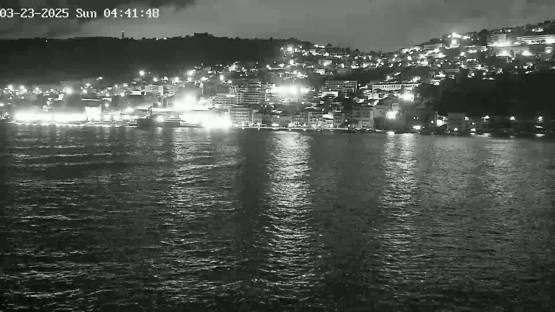 A nighttime, low-light view shows a city built on a hill, its lights reflected on a dark, moderately wavy body of water.
