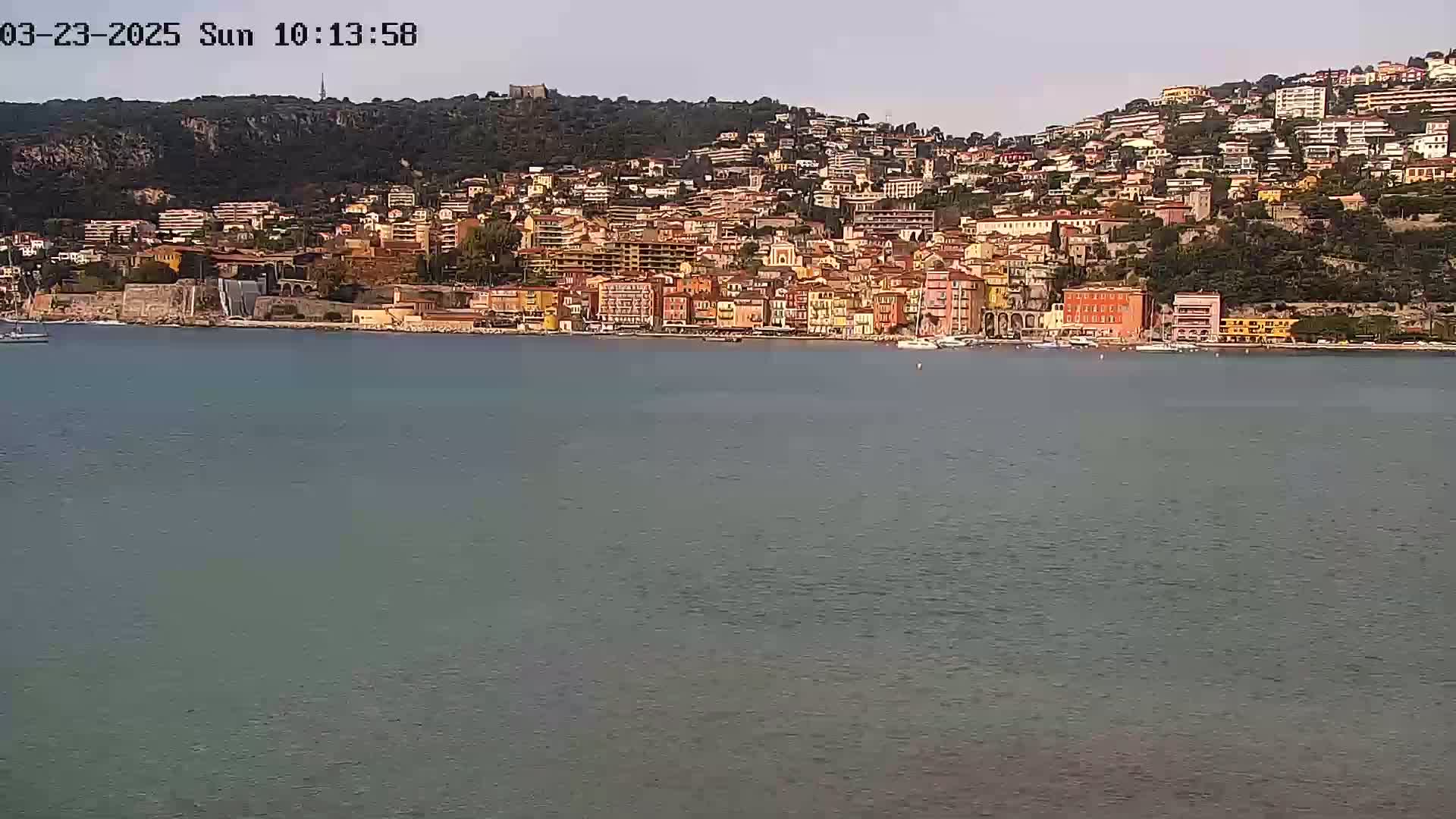 A calm body of water sits in front of a hillside town of closely packed colorful buildings under a clear sky.