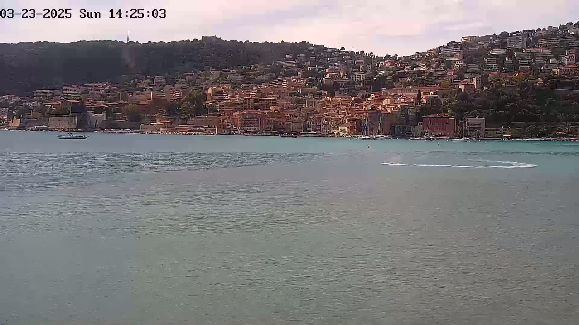 A coastal town with many red-roofed buildings climbs a hillside overlooking a calm body of water under a partly cloudy sky.