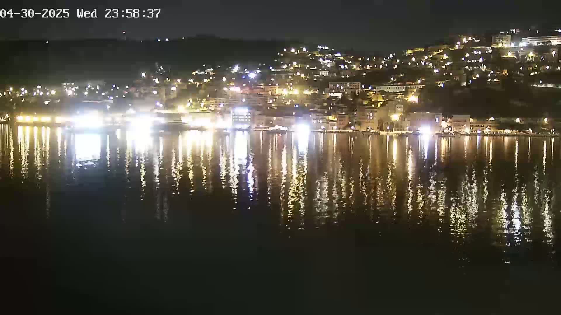 A nighttime view of a town situated on a hillside, its lights reflected in a calm body of water.