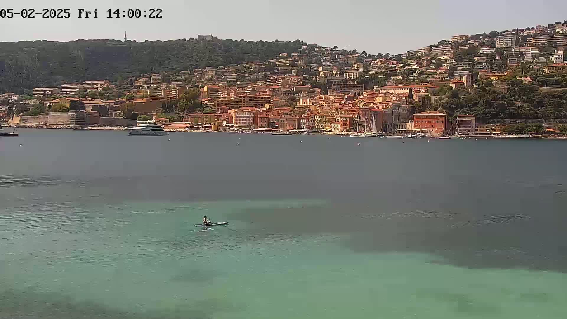 A person kayaks in calm, clear, teal water in front of a hillside town under a mostly clear sky.