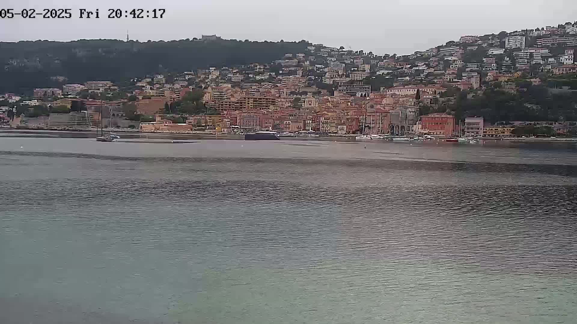 A hill covered in buildings sits on the far shore of a calm body of water under an overcast sky.