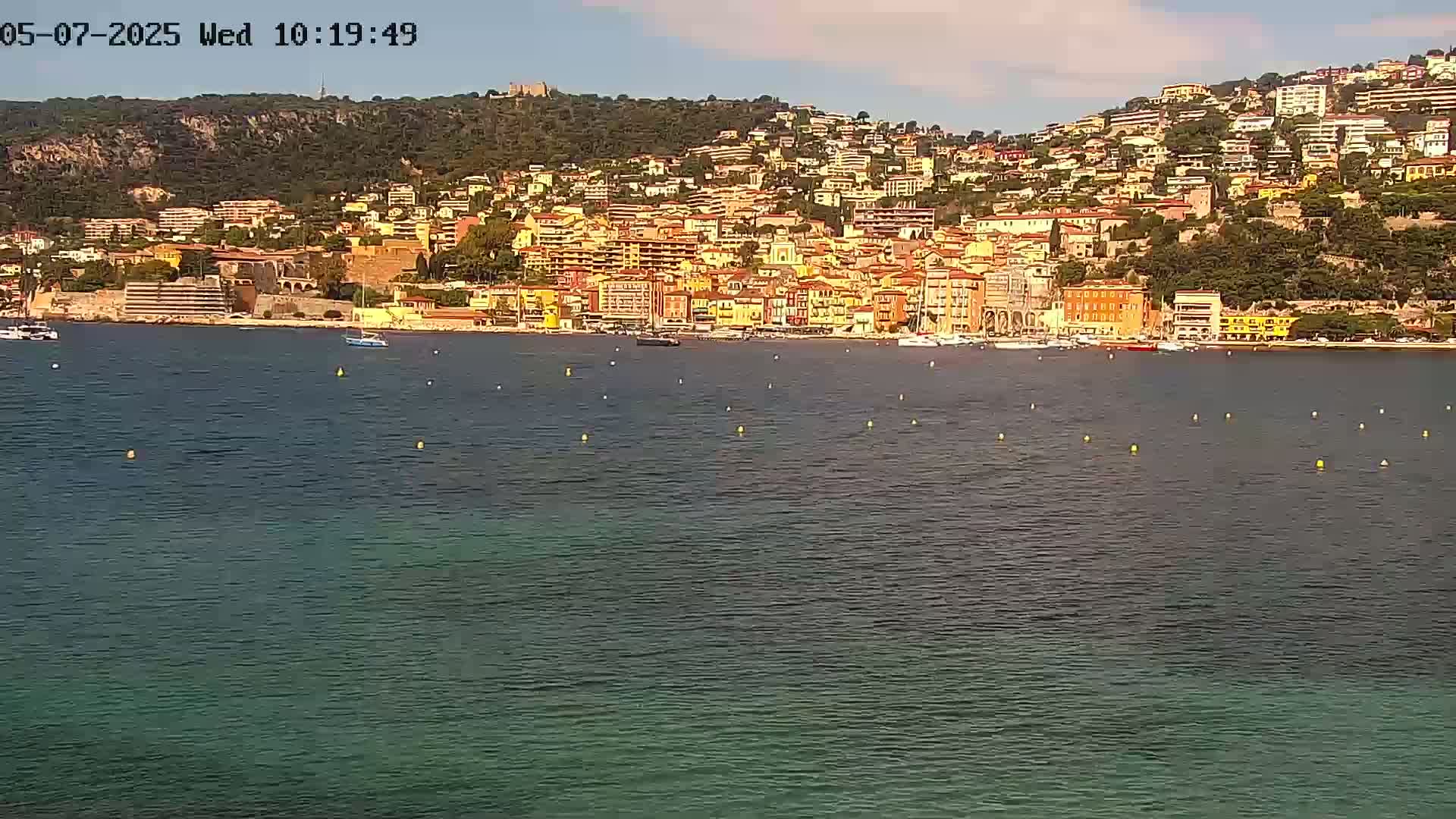 A coastal town with numerous buildings of varying heights and colors is situated on a hillside overlooking a calm body of water under a mostly clear sky.