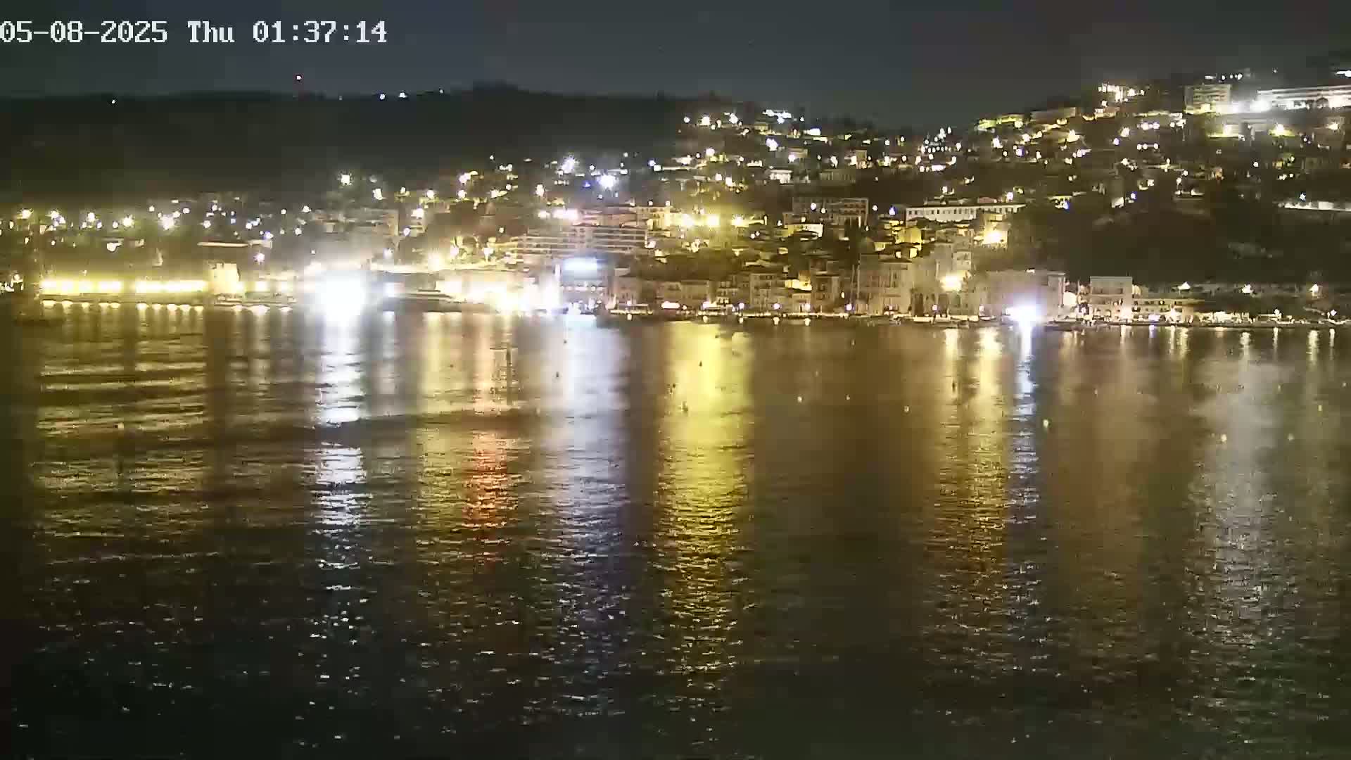 A nighttime view of a calm body of water reflecting the lights of a city built on a hillside.
