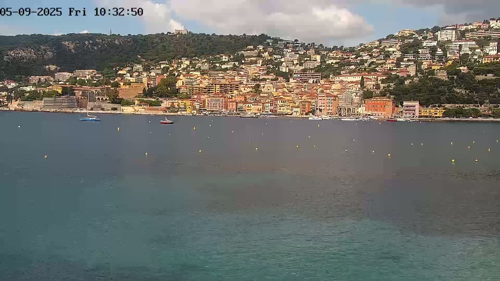 A picturesque coastal town, nestled against a hillside, is seen from across a calm body of water under a partly cloudy sky.