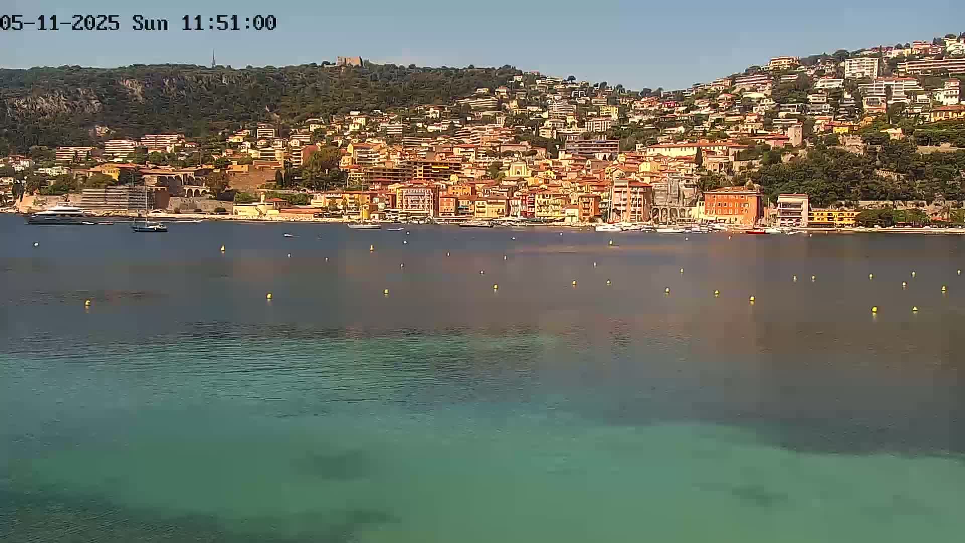 A hillside town of closely clustered buildings in warm, earth tones sits beside a calm, teal-colored body of water on a sunny day.