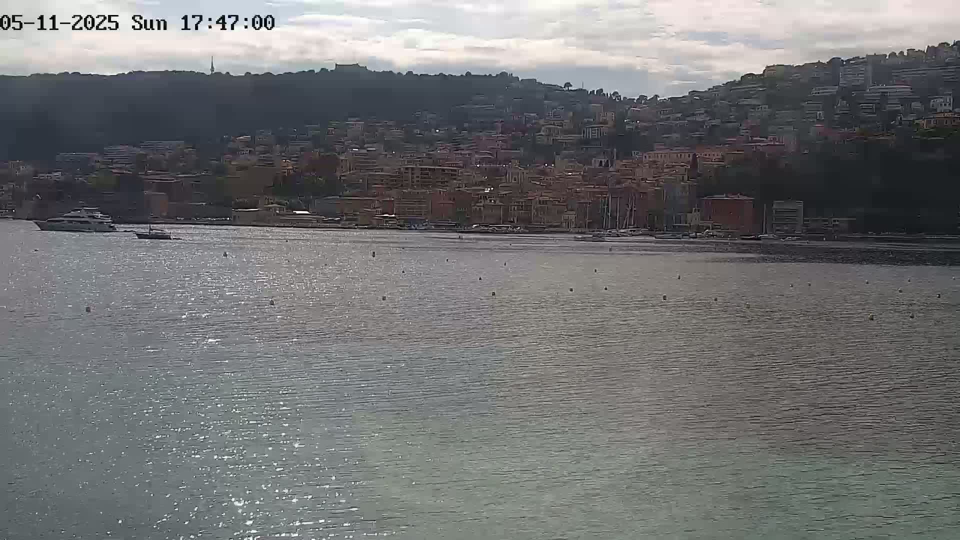 A calm body of water reflects the sunlight, with a cityscape of closely packed buildings on a hillside in the background under a mostly cloudy sky.