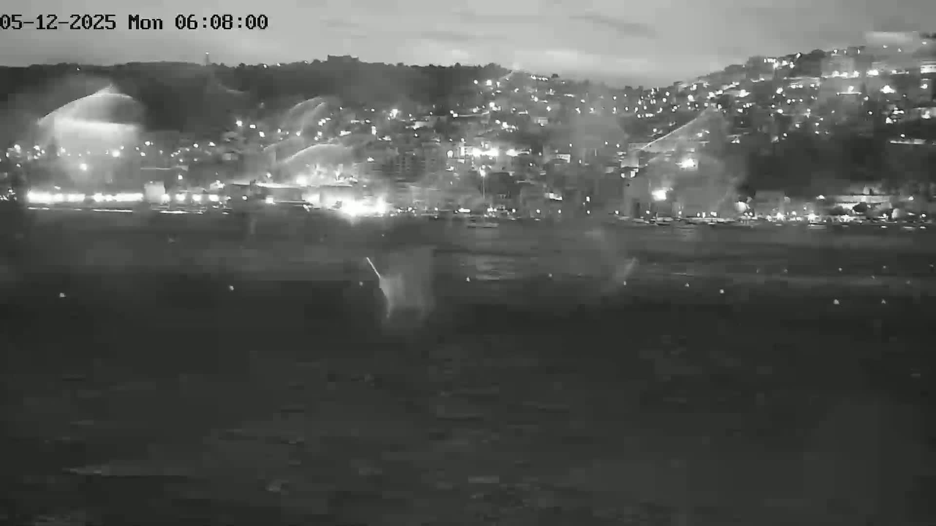 A nighttime, low-light view shows a town or city across a body of water, with some light rain or spray visible.