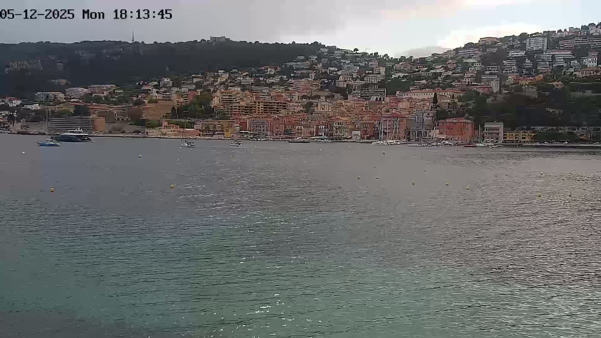 A waterfront town of multi-story buildings is nestled against a hillside under an overcast sky, with several boats in the calm water below.